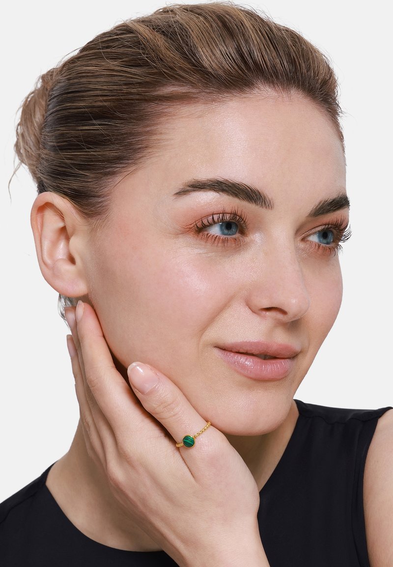 Bague en or ornée d'une pierre de malachite verte, avec un délicat anneau torsadé. Le modèle effleure légèrement son visage, portant un simple top noir.