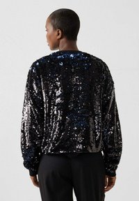 Persoon gezien van achteren, gekleed in een zwart-blauw top met lange mouwen en pailletten, en een zwarte broek, tegen een effen grijze achtergrond.