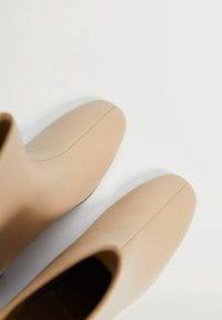 Botas de tobillo beige hechas de cuero suave con punta redondeada y detalles de costura mínimos. La suela muestra un contraste negro.