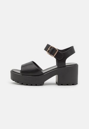 Sort chunky sandal med tyk sål, åben tå, ankelstropp og guldfarvet rektangulær spænde på stroppen.