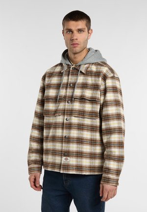 Ung man som bär en brun- och vitrutig knäppbar jacka över en grå huva och blå jeans, står mot en enfärgad bakgrund.