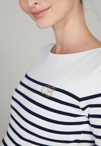 Wit en marineblauwe gestreepte t-shirt met een ronde hals, voorzien van een gouden geborduurd logo op de borst. Zacht, aansluitend materiaal.