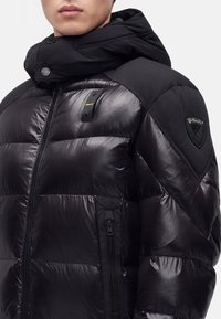 Homme portant une doudoune noire brillante avec capuche et poches zippées, munie d'une petite étiquette sur la poitrine et d'un patch logo sur l'épaule.
