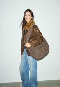 Kvinde iført en læderjakke med brun pelskrave, løse blå jeans, hvide sko og bærende en stor brun skuldertaske mod en ensfarvet væg.