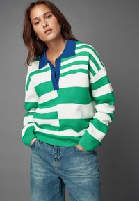 Grøn- og hvidstribet sweater med en blå krave, der har tre knapper. Blød tekstur, afslappet pasform, parret med lyseblå jeans.