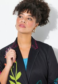 Schwarze Blazer mit farbenfroher Blumenstickerei und einem pinken gestickten Kragen. Das Model trägt ein blaues Oberteil darunter und goldene Creolen.