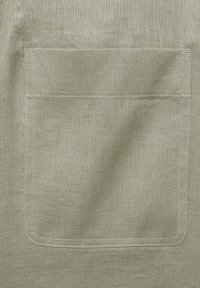 Svetlozeleni laneni material z velikim kvadratnim žepom. Tekstura izgleda rahlo nagubana, z vidnimi šivi ob robovih.