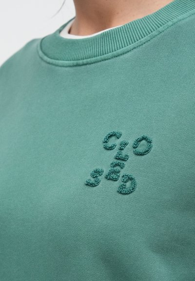 Κοντινό πλάνο ενός ατόμου που φοράει πράσινο φούτερ με στρογγυλή λαιμόκοψη, με υφασμένα, ανάγλυφα γράμματα "CLOSID" στο στήθος.