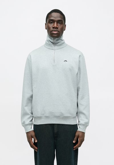 Homme noir portant un pull zippé gris clair avec un petit logo sur la poitrine, se tenant avec les mains détendues sur les côtés, sur fond blanc.