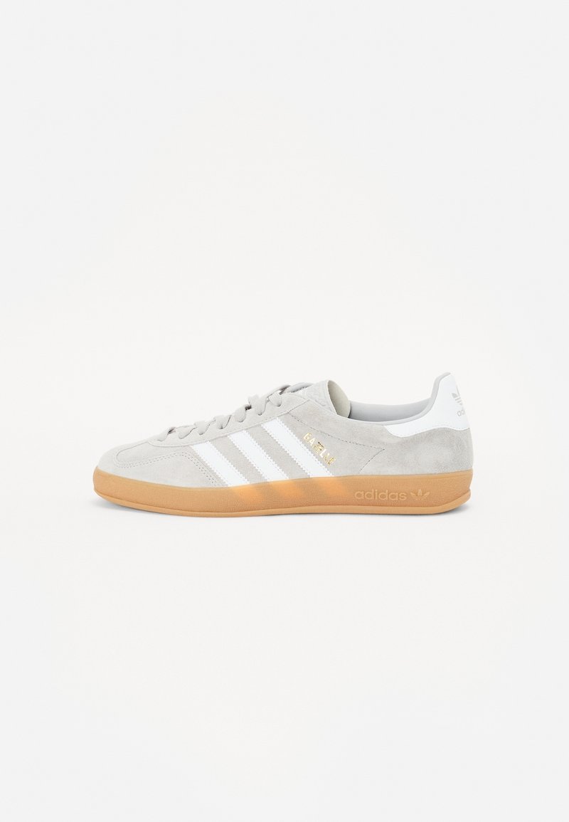 Szary zamszowy tenisówka z białymi skórzanymi paskami, gumową podeszwą w kolorze karmelowym i złotymi akcentami. Płaski, sznurowany design, zaokrąglony nosek, wyściełany kołnierz.