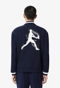 La chaqueta de marina presenta un gráfico de silueta atlética blanca en la parte posterior, con puños y cuello a rayas. Fabricada con un tejido suave, su diseño es casual.