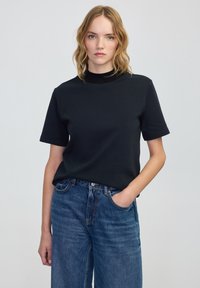 Černé tričko s krátkým rukávem a vysokým výstřihem, vyrobené z hladkého materiálu. V kombinaci s modrými volnými džíny, které mají uvolněný střih.