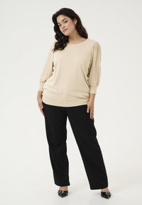 Beige gebreid topje met pofmouwen, gecombineerd met zwarte wijde broek en zwarte puntige schoenen, tegen een effen witte achtergrond.