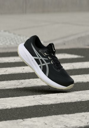 Pantof de alergare alb-negru, surprins în aer deasupra unei treceri de pietoni pe asfalt, cu mici resturi ridicate în urma tălpii.