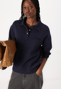 Donkerblauwe sweatshirt met kraag, knoopsluiting en geborduurd logo. Gecombineerd met grijze denim, met een glanzende bruine tas.