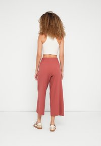 Femme aux cheveux bouclés se tenant face à un mur blanc, portant un crop top blanc, un pantalon ample couleur rouille et des baskets beiges.