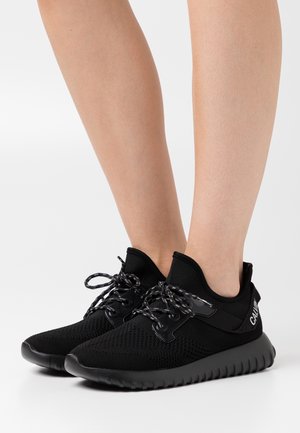 Personne portant des baskets basses en tricot noir avec des lacets à motifs et des semelles épaisses texturées sur fond blanc.