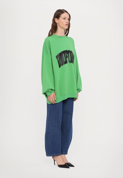 Grønn oversized hettegenser med svart grafisk tekst, kombinert med vide denimbukser og svarte spisse hæler. Glatt stoff og avslappet passform.