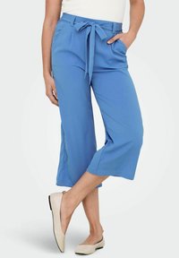Pantalon court bleu clair avec ceinture à nouer et poches latérales, en tissu texturé et coupe décontractée. Associé à des ballerines beiges.
