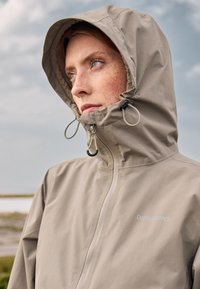 Woman with freckles wearing a beige rain jacket with hood up, looking into the distance against a cloudy sky background.