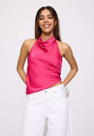 Usmievavá žena s rovnými vlasmi, ktorá nosí žiarivo ružový tielko bez rukávov a biele nohavice s vysokým pásom na jednoduchom svetlom pozadí.