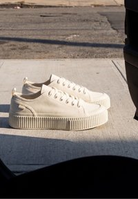 Paire de baskets compensées en toile couleur crème avec des semelles épaisses nervurées posées sur un trottoir en béton près d’un bord de rue.