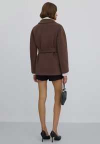 Brązowa kurtka z mieszanki wełny z paskiem w talii, dużym kołnierzem i przeszyciami, zestawiona z czarnymi szortami i butami na wysokim obcasie.