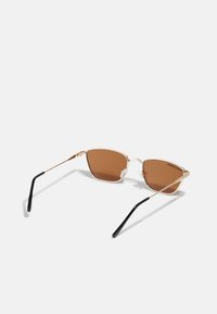 Ochelari de soare cu nuanța maro, având o ramă metalică aurie minimalistă. Lentilele sunt unghiulare, cu capace din plastic negru pe brațe pentru confort.