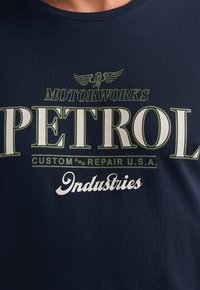 Tmavě modré tričko z měkké látky s velkým bílým a zeleným grafickým textem: "PETROL MOTORWORKS CUSTOM and REPAIR U.S.A." s motivem orla.
