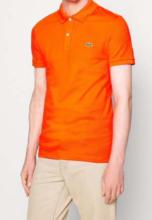 Oranges Poloshirt aus Baumwolle mit weicher Textur. Verfügt über einen Kragen, eine dreiknöpfige Blende und ein grünes Logo auf der Brust.