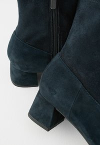 Mörkblå mocka ankelstövlar med klack och en sidodetalj med dragkedja, som har en texturerad yta och en strukturerad silhuett.