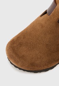 Braune Wildleder-Slipper mit abgerundetem Kopf, versehen mit einem dunklen Akzent und strukturierter Oberfläche. Enthält eine schwarze Gummisohle für besseren Halt.