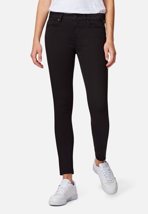 Jeans skinny negros con una textura suave y un diseño clásico de cinco bolsillos. Cuenta con cremallera y cierre de botón. Se llevan con zapatillas blancas.