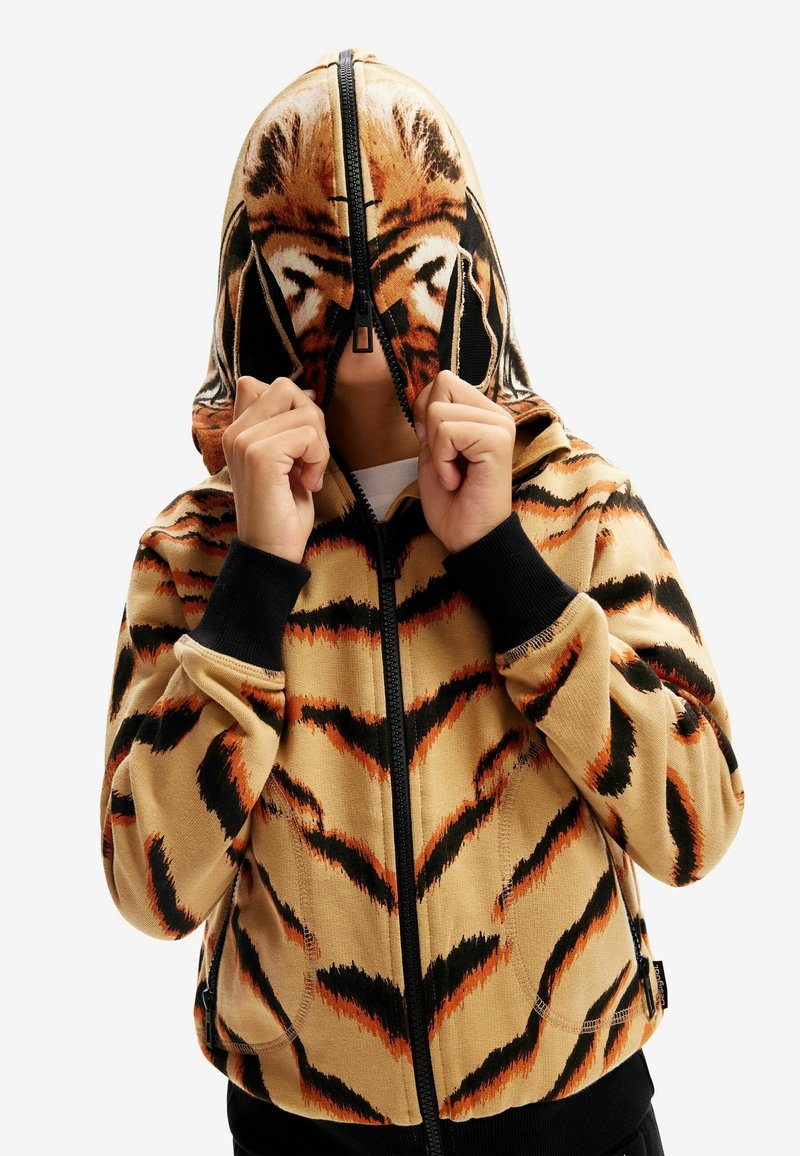 Hoodie zippé à motif tigre en beige avec des rayures orange et noires. Comprend une grande capuche et des poignets noirs côtelés. Texture lisse.