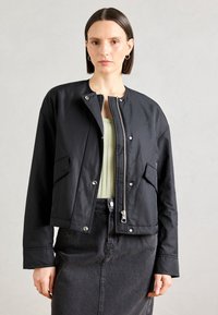 Černá bomber bunda s zipem vpředu, kovovými cvoky, dvěma bočními kapsami a zkráceným designem, spojená s světle zeleným topem a tmavou sukní.