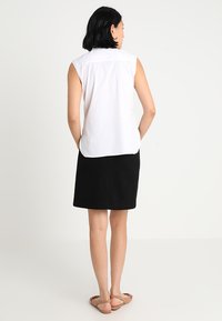 Blusa bianca senza maniche con colletto a punta abbinata a una gonna nera al ginocchio, ambientata su uno sfondo neutro. Sandali completano l'outfit.