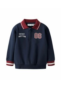 Tumesinine kampsun punase ja valge triibulise kraega. Omab valget tikandit tekstiga "VINTAGE RACE TEAM" ja numbri "86". Elastsed varrukaotsad.