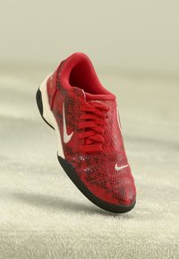 Sarkans sporta apģērbs ar melnu zoli, baltām Nike swoosh logo un auklu dizainu uz gaišas teksturētas virsmas.