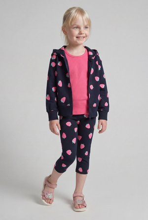 Lachend jong meisje in een marineblauwe hoodie en legging met patroon van roze aardbeien, draagt een roze shirt en roze sandalen met zilveren hartjes.