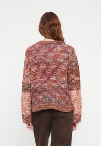 Strikkegenser i varme nyanser av oransje, maroon og beige. Løs passform med lange ermer og rund hals. Teksturert, lagdelt design.