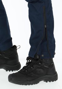 Zwarte wandelschoenen met getextureerde rubberen zolen en veters, gecombineerd met donkerblauwe broek met ritsen en drukknoopsluitingen op de enkels.