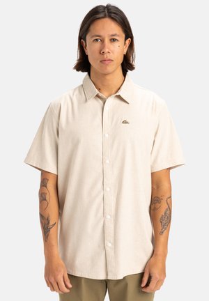 Beige Kurzarm-Hemd mit Knopfleiste und spitzem Kragen. Verfügt über ein kleines Logo und einen glatten, strukturierten Stoff. Getragen mit olivgrünen Hosen.