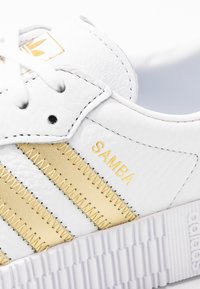 Primo piano di una sneaker Adidas Samba bianca con strisce beige, lacci bianchi e testo dorato "SAMBA" di lato.