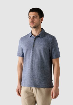 Hombre con una camisa polo de manga corta azul y pantalones beige, de pie contra un fondo liso gris claro, mirando hacia un lado.