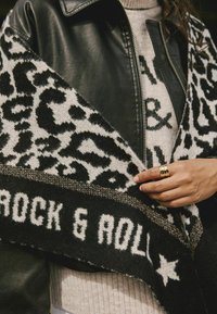 Sort læderjakke med et mønstret sort og hvidt strikket tørklæde med teksten "ROCK & ROLL." Iøjnefaldende guldring på hånden, struktureret materiale-detail.