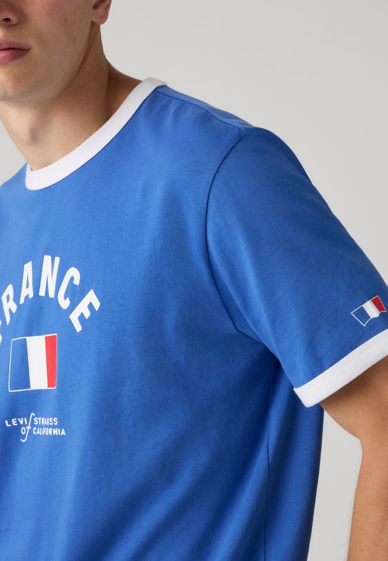 Uomo con maglietta blu con colletto bianco e polsini, con la scritta "FRANCE" e una grafica della bandiera francese davanti e sulla manica.