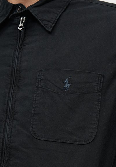 Μαύρο μπουφάν με γιακά και φερμουάρ μπροστά, με τσέπη στο στήθος που έχει κεντημένο λογότυπο παίκτη πόλο.