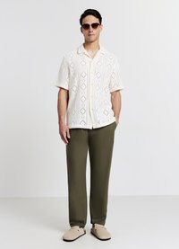 Weißes Strickshirt mit kurzen Ärmeln und Rautenmuster, olivgrüne Hose und beige Slipper. Mann mit runden Sonnenbrillen, steht aufrecht.