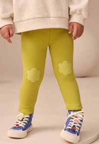 Grüne Leggings mit blumenförmigen Flicken an den Knien, kombiniert mit blauen Turnschuhen mit bunten Mustern und weißen Schnürsenkeln.