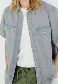 Camicia di jeans a maniche corte, azzurra, con bottoni a scatto, due tasche sul petto e una vestibilità comoda, indossata sopra una t-shirt bianca e pantaloni verdi.